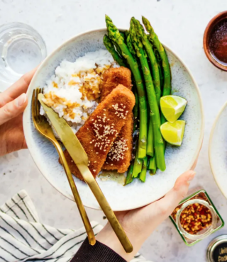 Eine Person hält einen Teller Kokosreis mit Spargel und Veggie Fisch von FRoSTA – panierter Tofu, Sesam, Sauce, Spargel und Limetten anbei.