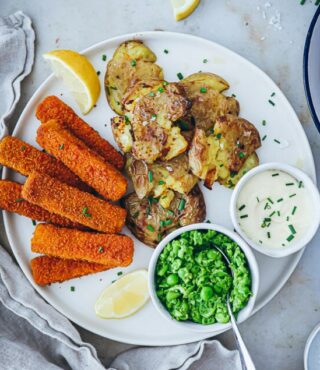 Ein weißer Teller mit FRoSTA Crispy Potatoes, FRoSTA Veggie Fisch, Kartoffelpüree, Zitronenspalte, Erbsen und Sahnesauce mit Schnittlauch. Gabel und Serviette liegen bereit.