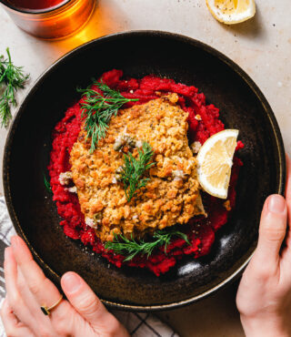 Eine Person hält eine schwarze Schüssel mit FRoSTA Veggie Schlemmerfilet und Rote-Bete-Meerrettich-Püree, garniert mit Dill und Zitrone.