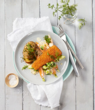 Ein Teller mit FRoSTA Veggie Fischfilet, Dill-Fächerkartoffeln und FRoSTA Gemüse, garniert mit frischem Dill, liegt auf einem weißen Holztisch.