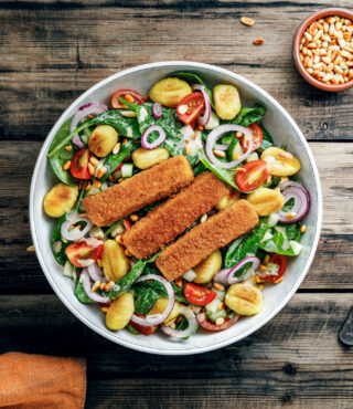 Eine Salatschüssel mit Spinat, Gnocchi, Kirschtomaten, roten Zwiebeln und drei FRoSTA Veggie Fischstäbchen obendrauf, Pinienkerne dazu.