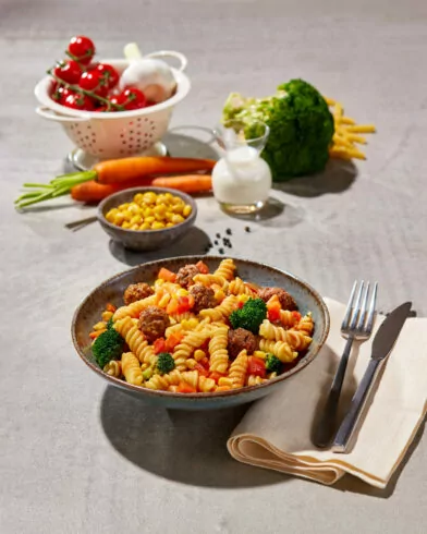 Eine Schüssel FRoSTA Rotini Pasta mit Brokkoli, Mais, Karotten und Hackbällchen steht auf dem Tisch – FRoSTA Gemüse und Milch dahinter.