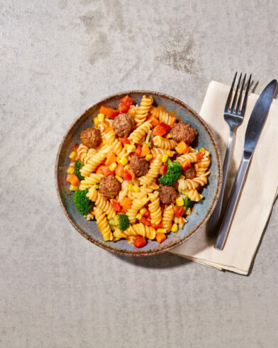 Eine Schüssel Fusilli-Nudeln mit Gemüse und kleinen Hackbällchen, wie aus einer klassischen FRoSTA Hackbällchenpfanne, serviert mit Besteck auf einer Serviette.
