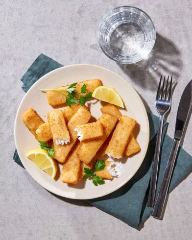 Ein Teller mit goldbraunen, knusprigen FRoSTA Fischstäbchen bio, garniert mit Zitrone und Petersilie, steht neben Wasser, Gabel und Messer auf einer grünen Serviette.