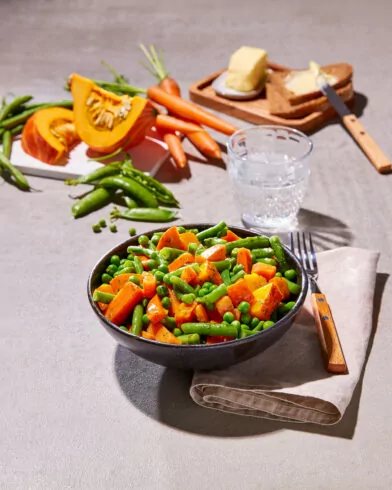 Eine Schüssel mit FRoSTA-Tiefkühlprodukten wie Karotten, grünen Bohnen und Erbsen steht auf einer Serviette, dazu frisches Gemüse im Hintergrund.