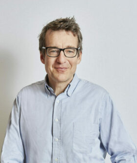 Ein Mann mittleren Alters mit braunen Haaren und Brille lächelt, im hellen Hemd vor neutralem Hintergrund – inspiriert von FRoSTA-Tiefkühlprodukten.