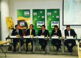 Fünf Männer in Anzügen sitzen am Konferenztisch, flankiert von grünen Bannern mit FRoSTA-Logo und FRoSTA-Tiefkühlprodukten nach Reinheitsgebot.