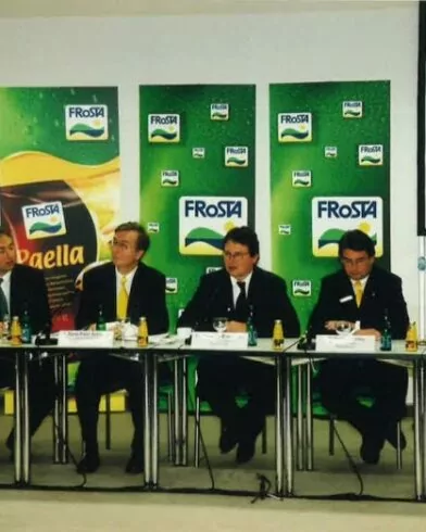 Fünf Männer in Anzügen sitzen am Konferenztisch, flankiert von grünen Bannern mit FRoSTA-Logo und FRoSTA-Tiefkühlprodukten nach Reinheitsgebot.