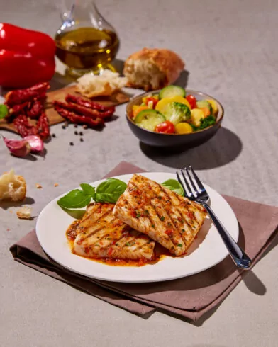 Zwei gegrillte FRoSTA Seelachsfilets mit Kräutern und Sauce auf einem weißen Teller, garniert mit Basilikum. Im Hintergrund Brot, Gemüse und FRoSTA Gemüse.