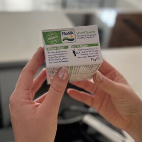 Eine Person mit hellrosa Nagellack hält eine kleine FRoSTA Kräuter-Packung Schnittlauch und zeigt Nährwerte sowie Umweltinfos auf Deutsch.