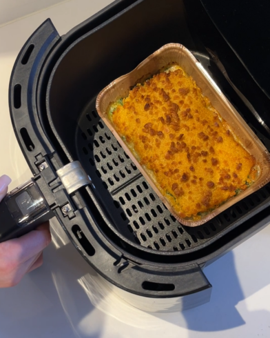 Eine Hand öffnet das Heißluftfritteusen-Fach und zeigt ein goldbraun gebackenes FRoSTA-Tiefkühlgericht in einer rechteckigen Aluschale.