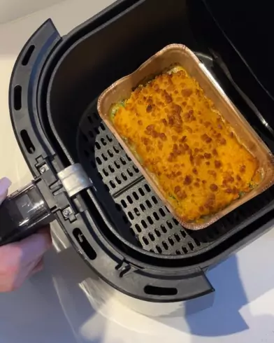 Eine Hand öffnet das Heißluftfritteusen-Fach und zeigt ein goldbraun gebackenes FRoSTA-Tiefkühlgericht in einer rechteckigen Aluschale.