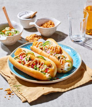 Zwei Hot Dogs mit frittierten FRoSTA-Tiefkühlprodukten, frischem Gemüse und Sauce auf einem blauen Teller, dazu Beilagen im Hintergrund.