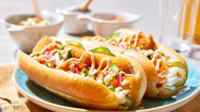 Zwei Hot Dog Brötchen mit knusprigem FRoSTA Fisch, Gurken, Zwiebeln und cremiger Sauce auf blauem Teller, im Hintergrund unscharfe Schalen.