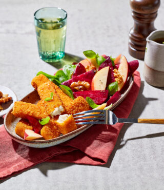 Ein Teller mit panierten Fischstäbchen von FRoSTA, Rote-Bete-Apfel-Salat, Walnüssen und Blattgemüse steht auf einer roten Serviette. Auf dem Teller liegt eine Gabel.