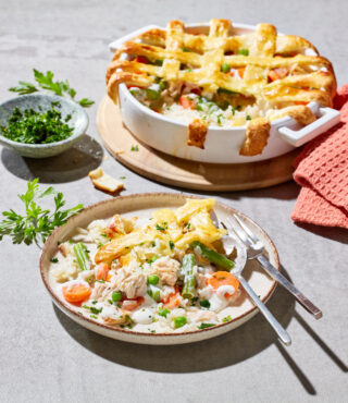 Eine Portion FRoSTA Chicken Pot Pie mit Gemüse und goldener Kruste liegt auf einem Teller, umgeben von frischen Kräutern und einem Tuch.