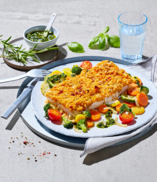 Ein Teller mit einem gebackenen FRoSTA Fischfilet in goldbrauner Kruste, serviert mit FRoSTA Gemüse und grüner Sauce. Gabel, Messer, Wasserglas und Kräuter liegen bereit.
