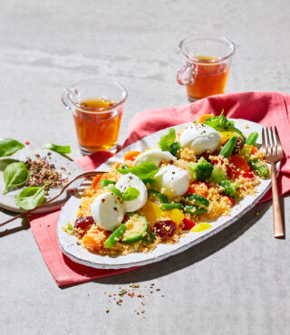 Ein weißer ovaler Teller mit einem Salat aus FRoSTA Quinoa, Kirschtomaten, Gurken, Paprika, Basilikumblättern und Mozzarella-Kugeln. In der Nähe: Goldgabel, Basilikumblätter, FRoSTA Quinoa und zwei Gläser Tee auf roter Serviette.