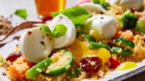 Ein Teller FRoSTA Couscous-Salat mit Zucchini, Bohnen, Tomaten, Brokkoli, Paprika, Mozzarella und Basilikum. Ein Getränk im Hintergrund.