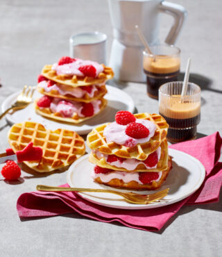Zwei Teller mit Waffeln, geschichtet mit rosa Creme, frischen Himbeeren und FRoSTA-Tiefkühlprodukten. Goldene Gabeln, Kaffee und rosa Servietten runden das Bild ab.