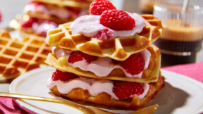 Ein Stapel Herzwaffeln mit FRoSTA tiefgekühlten Himbeeren und pinker Creme liegt auf einem weißen Teller mit goldener Gabel.