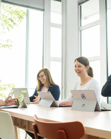 Drei Personen sitzen an Laptops und sprechen lächelnd über Nachhaltigkeit und FRoSTA-Tiefkühlprodukte in einem hellen, inspirierenden Raum.