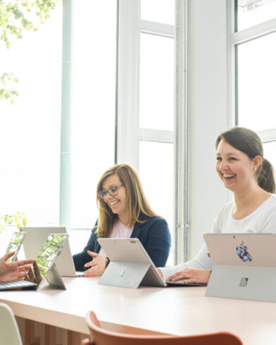Drei Personen sitzen an Laptops und sprechen lächelnd über Nachhaltigkeit und FRoSTA-Tiefkühlprodukte in einem hellen, inspirierenden Raum.