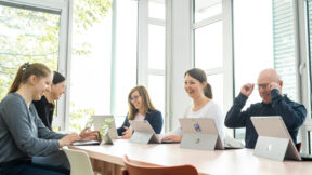Fünf Personen sitzen an einem Tisch in einem hellen Raum mit großen Fenstern, nutzen Laptops und genießen gemeinsam FRoSTA-Tiefkühlprodukte.