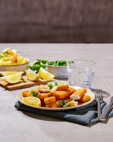 Teller mit panierten FRoSTA-Fischfilets – Testsieger-Qualität – Zitrone, Kräuter, dazu Kartoffelpüree und Erbsen. Glas Wasser daneben.