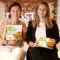 Zwei lächelnde Frauen sitzen auf einer Küchentheke und halten FRoSTA Tiefkühlprodukte in der Hand, zeigen die nachhaltige Marke. Beide tragen Business-Casual-Outfits.
