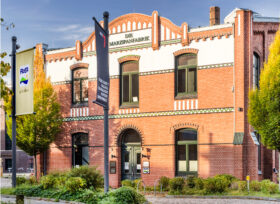 Ein historisches rotes Backsteingebäude mit der Aufschrift DIE MARZIPANFABRIK steht als Symbol für Nachhaltigkeit – mit FRoSTA-Tiefkühlprodukten und Herbstbäumen davor.