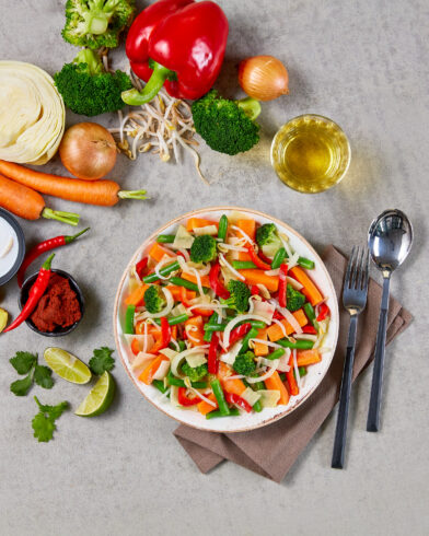 Neben einer Serviette steht eine Schüssel Salat mit Karotten, Paprika und Gemüse. Daneben rohe FRoSTA Bio Gemüse Mix Zutaten nach Thai Art: Karotte, Kohl, Chili, Zwiebel, Limette.