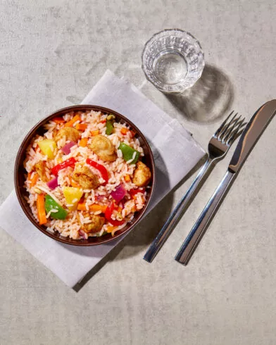 Eine Schale Reis mit Gemüse und FRoSTA Hähnchen Süß Sauer-Bällchen steht neben Wasser, Gabel und Messer auf heller Oberfläche.