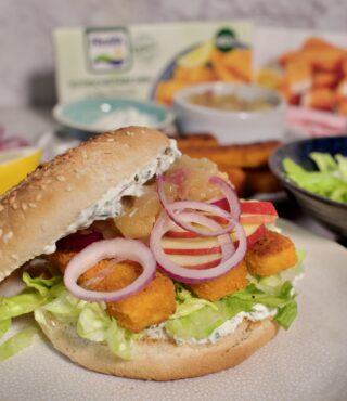 Ein Sesam-Sandwichbrötchen mit Salat, panierten FRoSTA Fischburger-Sticks, Tomaten, roten Zwiebeln und heller Sauce. Im Hintergrund die FRoSTA Verpackung.
