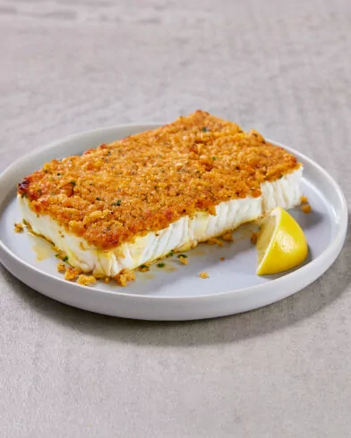 Ein FRoSTA Schlemmerfilet à la Bordelaise mit goldenem, knuspriger Kruste liegt neben einer Zitronenscheibe auf weißem Teller.