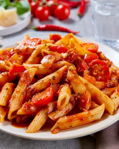 Ein Teller FRoSTA Tiefkühl-Bio Penne all'Arrabbiata mit Tomatensauce und Kirschtomaten steht neben Besteck und Wasser auf dem Tisch.