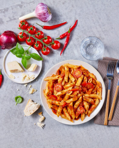 Eine Schüssel FRoSTA Bio Penne all'Arrabbiata mit Tomatensauce, Kirschtomaten und frischen Zutaten steht auf einer grauen Oberfläche.