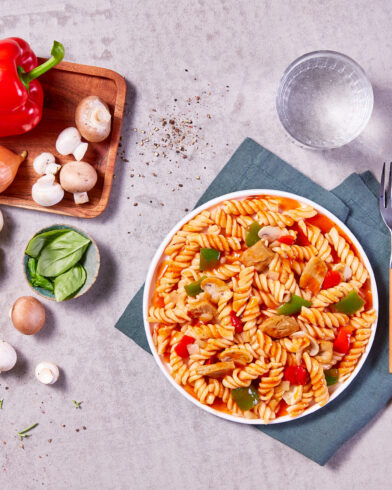 Auf einem Teller liegen Fusilli mit Tomatensoße, Paprika & Pilzen. Daneben Zutaten für FRoSTA Vegane Gulasch Pfanne auf dem Brett.