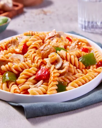 Ein Teller Rotini mit Tomatensauce, Hühnchen, Paprika und Pilzen erinnert an die FRoSTA Vegane Gulasch Pfanne. Mit Besteck und Wasser serviert.