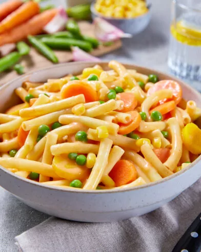 Eine Schüssel FRoSTA Bio Makkaroni Pfanne mit Karotten und Erbsen steht neben Besteck; im Hintergrund frisches Gemüse und ein Glas Wasser.