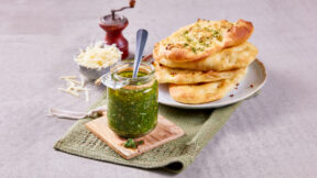 Glas schnelles Pesto mit Löffel auf Brett, dazu geriebener Käse, Pfeffermühle und FRoSTA-Tiefkühlprodukte auf grünem Tuch.