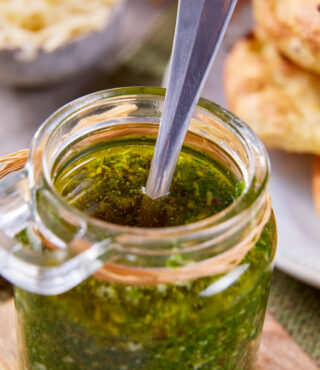 Ein Glas schnelles Pesto mit Löffel steht auf einem Brett. Dahinter FRoSTA-Tiefkühlprodukte, Fladenbrot und geriebener Käse.
