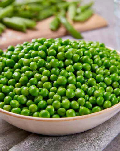Auf einem grauen Tuch steht eine Schüssel mit frischem FRoSTA Bioland Junge Erbsen, dahinter sind verschwommen ein Glas Wasser und Erbsen in ihren Schoten zu sehen.