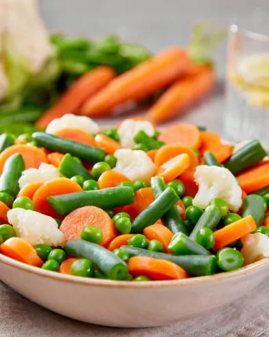 Eine Schüssel mit FRoSTA Tiefkühlprodukten – Bioland Sommergemüse mit Karotten, Erbsen, Bohnen & Blumenkohl – steht neben Wasser mit Zitrone.