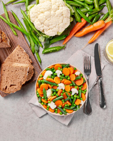 Eine Schüssel FRoSTA Tiefkühlgemüse – Karotten, Blumenkohl, grüne Bohnen, Erbsen – neben Zitronenwasser und Brot mit Butter auf einem hellgrauen Untergrund.