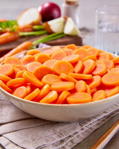 Eine Schüssel mit Bioland Karotten von FRoSTA steht auf einer Serviette, daneben ganze Karotten, Zwiebeln, Wasser und Besteck auf dem Tisch.