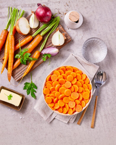 Draufsicht auf Bioland-Karotten, frische Zwiebeln, Petersilie und FRoSTA-Tiefkühlprodukte auf einem Schneidebrett, daneben geschnittene Karotten und Butter.