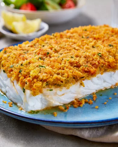 Ein gebackenes FRoSTA Schlemmerfilet mit Bio-Knusper-Auflage auf blauem Teller, dazu Zitronenspalten, Brot und Salat im Hintergrund.