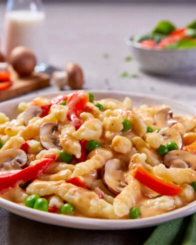 Ein Teller cremiger FRoSTA Bio Rahm Spätzle Pfanne mit Pilzen, Paprika und Erbsen steht auf dem Tisch, dazu Wasser und frisches Gemüse.