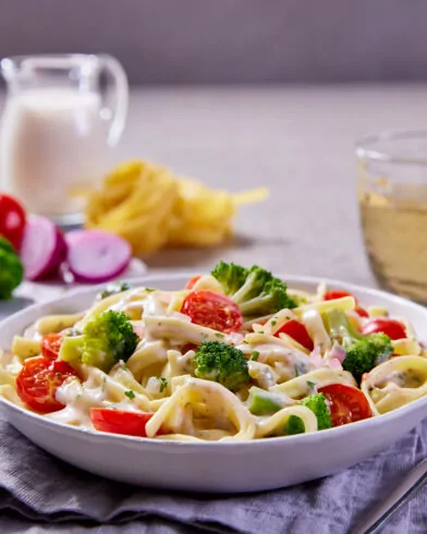 Eine Schüssel cremige FRoSTA Bio Pasta alla Toscana mit Brokkoli und Tomaten, dazu Weißwein, Brot und Gemüse auf dem Tisch.
